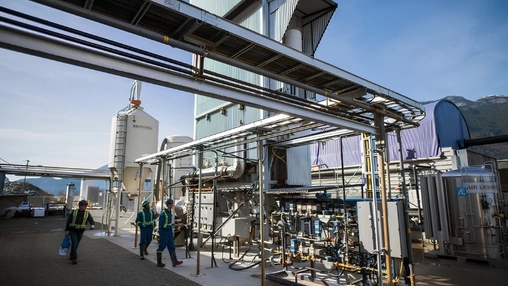 Outdoor industrial plant with workers in hi-vis gear walking past pipework and a cylindrical storage tank