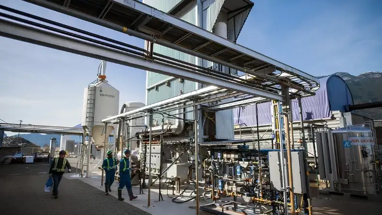 Outdoor industrial plant with workers in hi-vis gear walking past pipework and a cylindrical storage tank